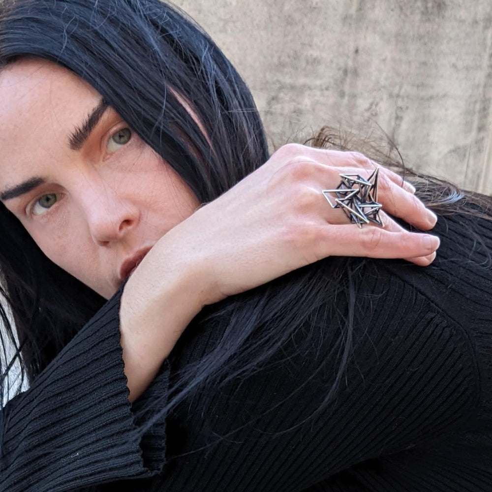
                  
                    A woman with long black hair wears a unique ring and rests her arm on her shoulder, dressed in a black ribbed top against a concrete background.
                  
                