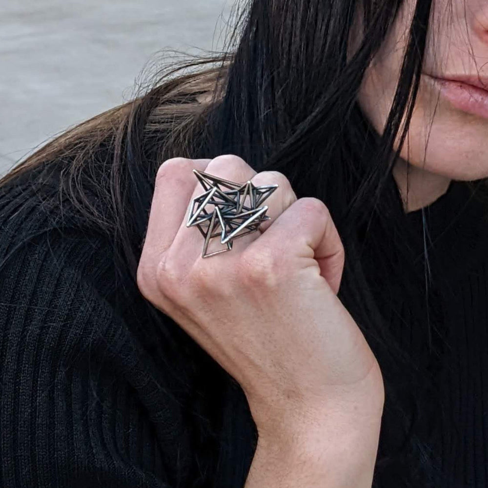 
                  
                    A person with long dark hair holds their hand up, showcasing a unique silver star-shaped ring against a textured concrete background.
                  
                