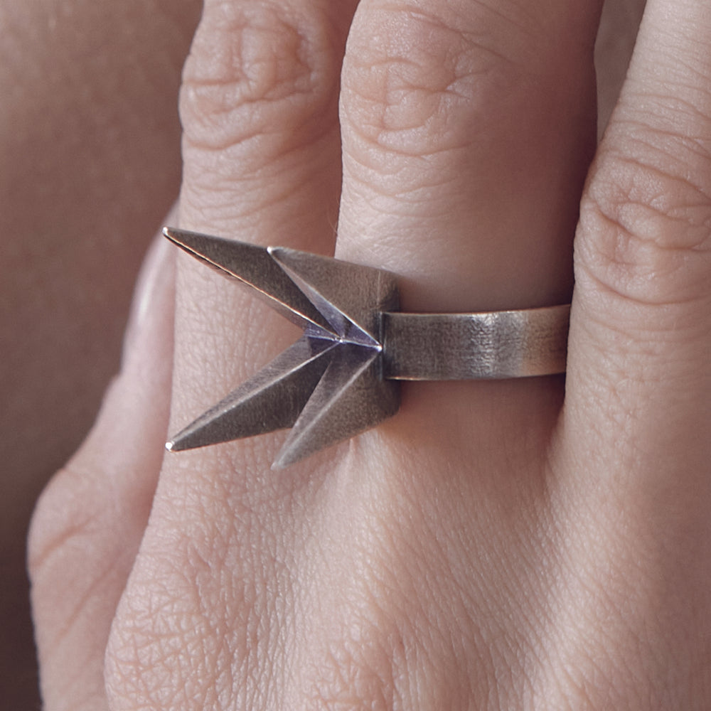 
                  
                    A close-up of a hand displaying a unique origami-inspired silver ring with pointed tips, resting against a neutral background.
                  
                