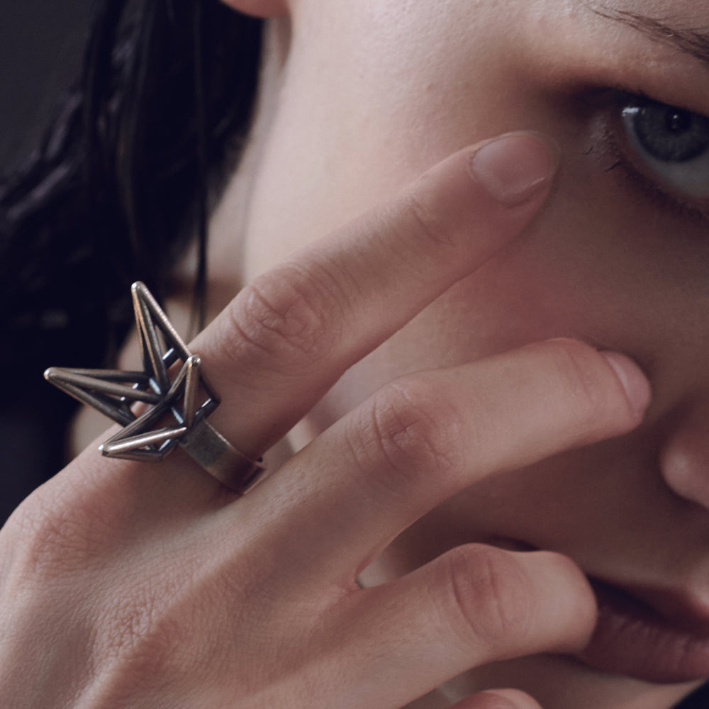 A close-up of a hand with a unique, metallic origami-inspired ring, gently touching the model's face. The model has youthful features.