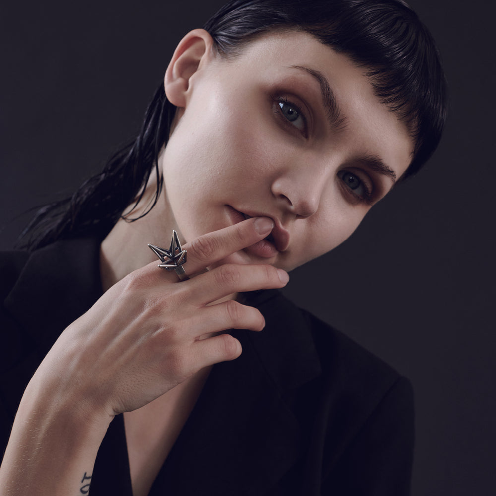 
                  
                    A close-up of a person wearing a dark blazer, with slicked-back hair and a hand holding a silver ring, set against a dark background.
                  
                